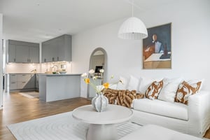 Ljust och modernt vardagsrum med vit soffa, prydnadskuddar och ett runt soffbord. Köksdel i grått med öppen planlösning samt en vas med blommor och en tavla på väggen