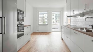 Modernt kök med vita skåp, inbyggd ugn och mikrovågsugn, träparkettgolv samt utsikt mot en innergård genom ett stort fönster och altandörr