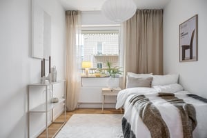 Sovrum med vit inredning, stor fönster med beige gardiner, säng med vit och grå bäddning, vit taklampa och dekorativ hylla