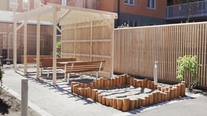 Lekplats med sandlåda omgiven av trästockar, sittplatser under tak och trädgårdsmiljö mot bakgrund av flerfamiljshus