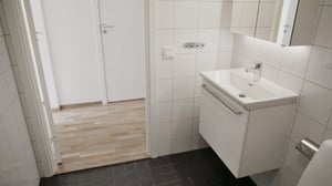 Ljust badrum med vita kakelväggar, vit kommod med spegel och svart klinkergolv, samt en öppning mot ett rum med parkettgolv