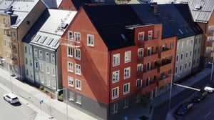 Flerbostadshus med fasader i olika färger, inklusive rött och grått, vid en gata med parkerade bilar och träd