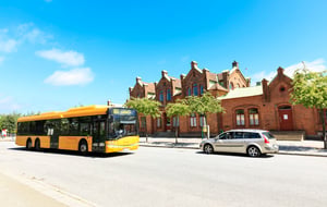 Gul stadsbuss och silverfärgad bil framför en röd tegelbyggnad under klarblå himmel
