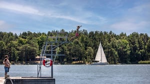 Person som hoppar från ett hopptorn vid en sjö, med segelbåt och skog i bakgrunden