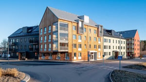 Flervåningshus i blandad arkitektur med trä- och betongfasad, beläget vid en korsning under klarblå himmel