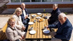 Fem äldre personer sitter vid ett utomhusbord med kaffe och fikabröd i soligt gårdsläge