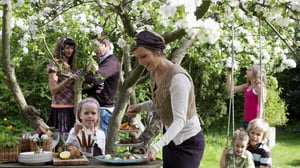 Flera vuxna och barn vid ett trädgårdsbord med serverad mat under blommande träd, utomhus i grönskan