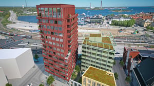 Två moderna byggnader, en röd och en grön, med gröna tak i ett stadslandskap nära vatten och hamn