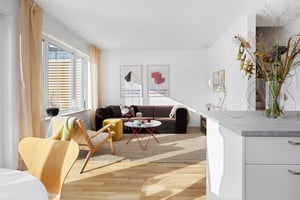 Ljus och modern vardagsrumsinteriör med stora fönster, en brun soffa, färgglada konstverk på väggen och en matplats med trästolar