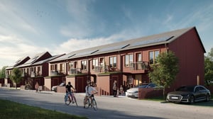 Rad av moderna röda radhus med trädetaljer, solpaneler på taket och omgivande grönska; människor och cyklister på gatan framför husen