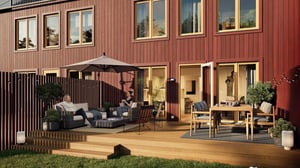 Radhus med röd fasad och stora fönster, en träaltan med möbler, parasoll och grönskande trädgård