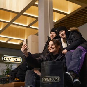 Personer tar en selfie i ett köpcentrum, med varm belysning och en tydlig CENTRUM-skylt i bakgrunden