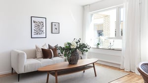 Ljust vardagsrum med soffa, soffbord, blommor och stora fönster i en inflyttningsklar lägenhet