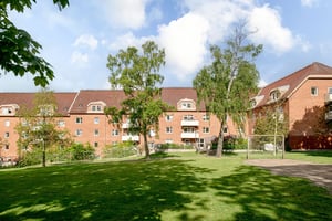 Flerfamiljshus i tegelfasad med takkupor och vita balkonger, omgiven av gräsmatta, träd och en liten fotbollsplan