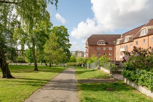 Flerfamiljshus i tegelfasad med takkupor och vita balkonger, omgiven av gräsmatta, träd och en stenbelagd gångbana