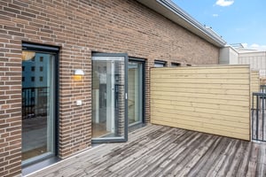 Balkong med trätrall, tegelvägg, glasdörrar och träpanel, omgiven av byggnader under klarblå himmel