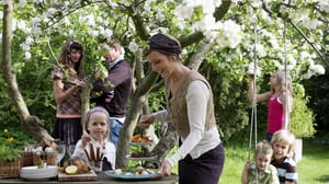 Flera vuxna och barn vid ett trädgårdsbord med serverad mat under blommande träd, utomhus i grönskan