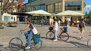 Människor cyklar och går framför Lunds stadshall med uteservering och blomsterarrangemang på torget