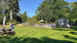 Personer sitter vid picknickbord i en grön park med uteservering och träd i bakgrunden