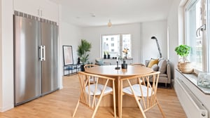 Inflyttningsklar lägenhet med ljust kök och matplats, stort kylskåp, runda matbord och fåtölj vid fönstret