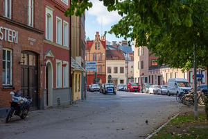 Gata i stadsmiljö med parkerade bilar, cyklar och byggnader i bakgrunden. Grönskande träd synliga på höger sida