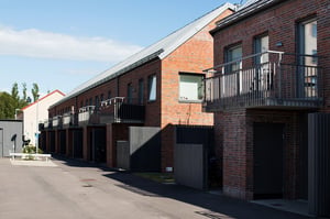 Radhus med fasad av rött tegel, balkonger i metall och svarta grindar, beläget vid en asfalterad väg under klarblå himmel