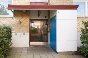 Ingång till ett flerfamiljshus med tegelvägg, blå dörr och glasad entréport