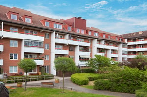 Flerfamiljshus med röda tak och vit-röda fasader, omgivna av grönskande trädgård och gångvägar