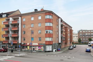 Fyravånings bostadshus i tegel med balkonger, butik på bottenvåningen och parkerade bilar längs gatan