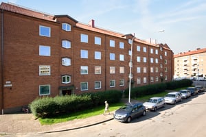 Gatvy med flerbostadshus i röd tegel, flera parkerade bilar och en person som går på trottoaren framför byggnaden
