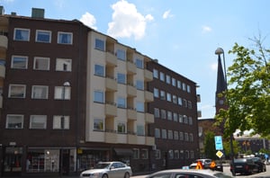 Flerbostadshus i brun och beige fasad med parkerade bilar på gatan, en kyrkobyggnad synlig i bakgrunden under en klarblå himmel