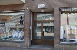 Entré till en byggnad med skylten "Akelius" ovanför dörren, omgiven av butiker med skyltfönster som innehåller konst och ramar
