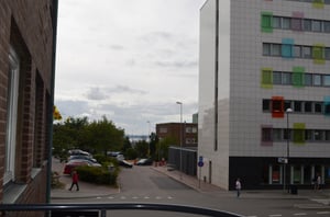 Vy över en stadsgata med parkerade bilar, träd och ett modernt byggnadskomplex med färgglada fönster. En person promenerar på trottoaren