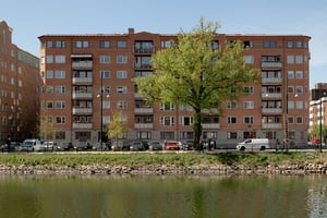 Flerfamiljshus i tegel och ljusa balkonger, omgiven av grönska och gata med vattenkanal i förgrunden