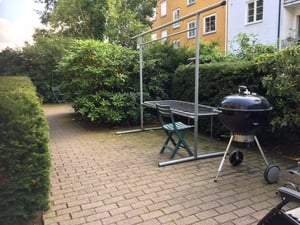 Grill och sittplats på en stenlagd uteplats omgiven av gröna buskar och träd, med ett gulmålat flerfamiljshus i bakgrunden