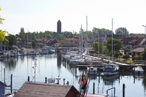 Småbåtshamn med segelbåtar och motorbåtar vid kaj, omgivna av hus och grönska med en kyrktornliknande byggnad i bakgrunden