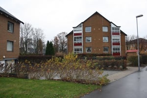 Brun tegelbyggnad med flera våningar, omgiven av buskar, trottoar och gräsmatta på en molnig dag