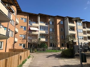 Flerfamiljshus i tegel med balkonger, grönområde och staket framför, under en klarblå himmel