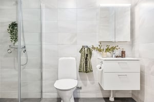Modernt badrum med vit inredning, toalett, dusch med glasvägg och spegelskåp ovanför ett vitt tvättställ. Gröna växter och en randig handduk tillför färg