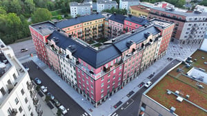 Flygbild av ett V-format färgglatt bostadskomplex med rosa, gula och beige fasader, omgiven av gator, byggnader och grönska