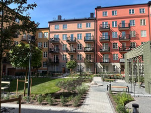 Flerfamiljshus med färgglada fasader, balkonger och en grön innergård med gångvägar och planteringar under en klarblå himmel