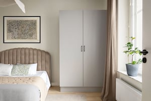 Sovrum med en beige sänggavel, vit sängklädsel, ljusgrå garderob och ett fönster med beige gardin. En grön växt i blå kruka står på fönsterbrädan