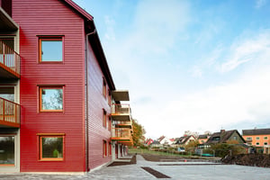 Flerfamiljshus med röd träfasad, balkonger och omgivande trädgårdar, med hus och blå himmel i bakgrunden