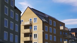 Gul byggnad med balkonger och svarta tak, omgiven av andra byggnader under en klar blå himmel