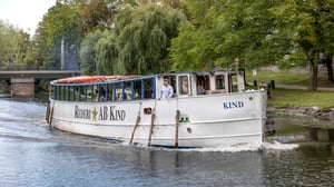En vit passagerarbåt med texten Rederi AB Kind på sidan som färdas på en flod omgiven av grönskande träd