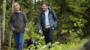 Två män promenerar med en svart hund i grön skogsmiljö