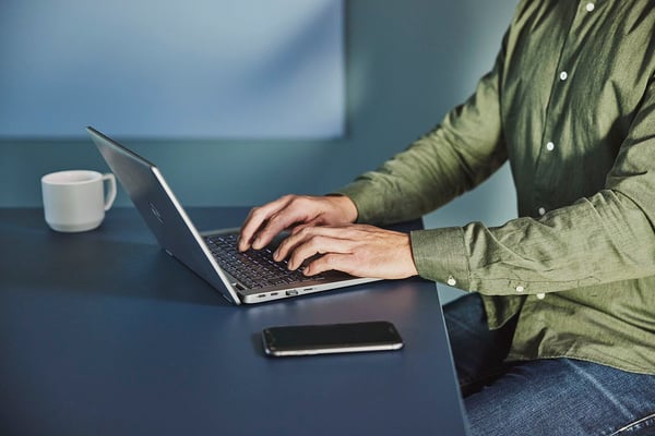 Man som sitter vid ett bord och skriver på en bärbar dator; en mobiltelefon och en kaffekopp syns bredvid