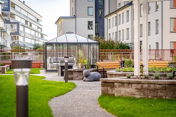 Grönyta med gångstig, stenmur, växter och ett växthus i en modern bostadsområde