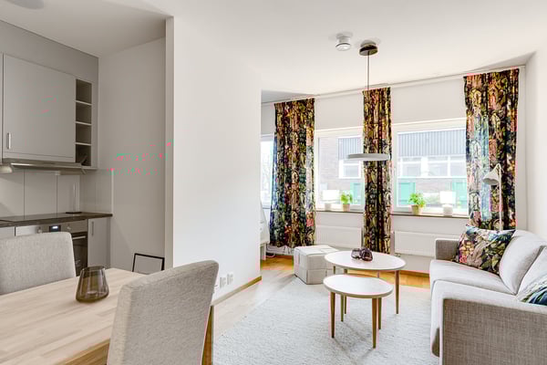 Ljust vardagsrum med soffa, soffbord och matbord, dekorerat med färgglada blommiga gardiner, bredvid ett kök med grå skåp