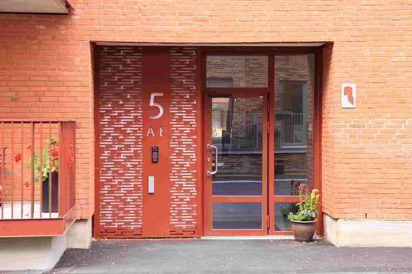 Entré till flerfamiljshus med en röd dörr och glasfönster, omgivna av tegelväggar. En krukväxt står vid sidan av dörren